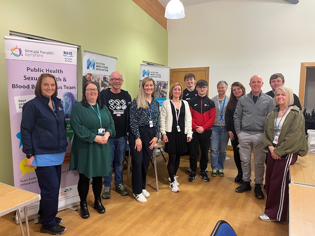 Staff from NHS Ayrshire & Arran and partners standing in front of information stall at Mens Health event.