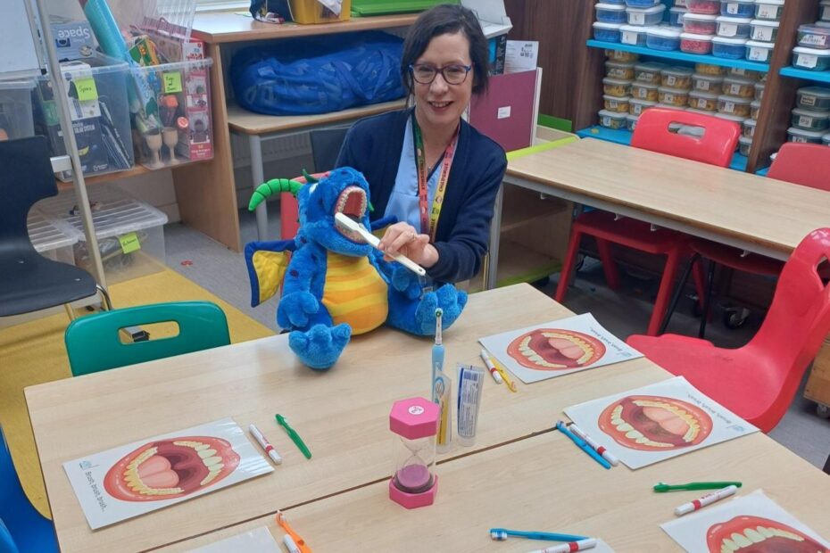 A member of NHS Ayrshire & Arran Oral Health team sitting in a primary school classroom surrounded by activities and holding a toy and toothbrush
