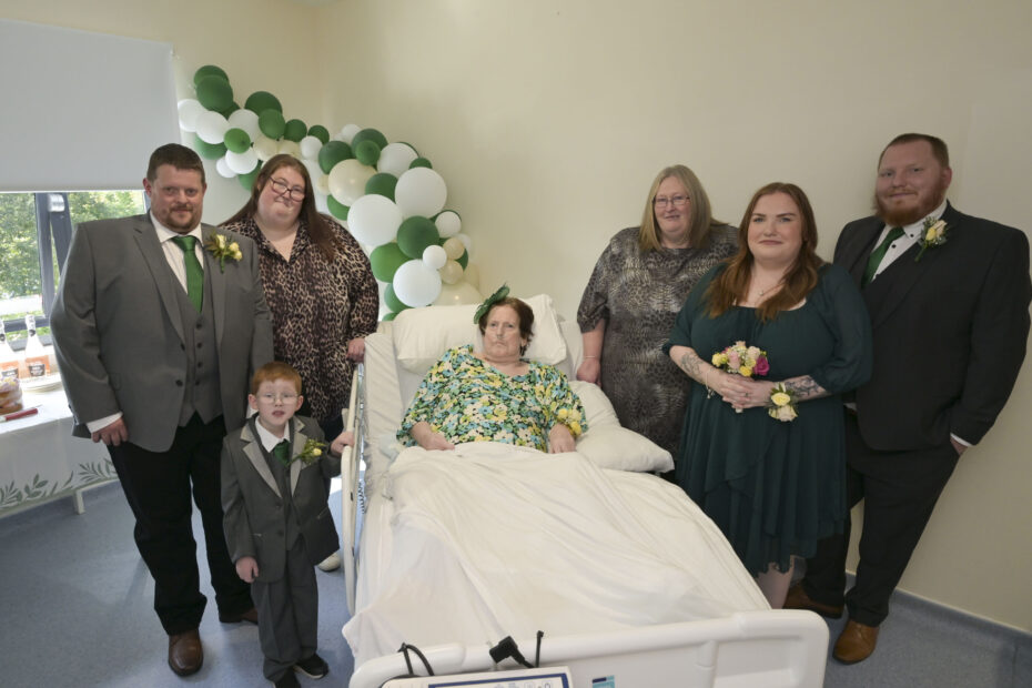 Hospital room, with Grooms mother in her ward bed, balloon arch in background. and from left to right James (groom), Shoni (bride), mum Carol and rest of family dressed for attending their wedding